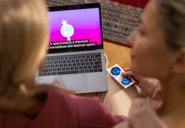 Two women watching an educational video on contraception and holding condoms indoors.