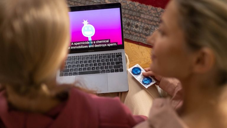 Two women watching an educational video on contraception and holding condoms indoors.